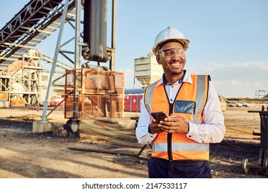 A young African mine worker wearing protective wear is looking off camera while holding a cell phone with coal mine equipment in the background