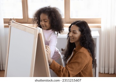 Young African mother her little adorable daughter drawing pictures chalkboard spend leisure in cozy modern nursery  Art class for preschool children  kids development  family creative hobby concept