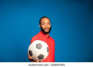 Young African Nigerian Man In Casual Red Shirt Posing Isolated On Blue Background Studio Portrait. People Lifestyle Concept. Holding Football Showing In To The Camera