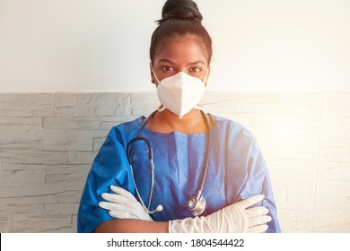 A Young African Nurse Wears A Blue Uniform, A Mask And Gloves While Standing With Her Arms Crossed In The Hospital.