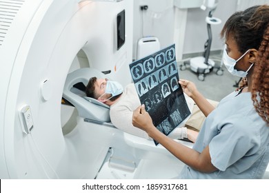 Young African Radiologist In Uniform And Protective Mask Looking At X-ray Image Of Patient Lying On Long Couchette Of Medical Equipment