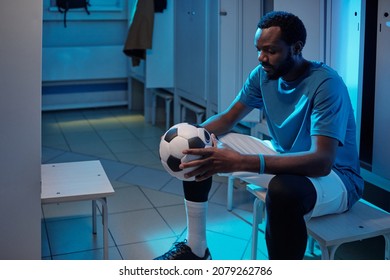 Young African sportsman looking at soccer ball in his hands while sitting by locker in changing-room at break - Powered by Shutterstock