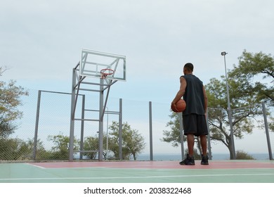 Young african sportsman standing with ball and looking on basketball hoop on sports court. Back bottom view of black man with towel wear sportswear and sneakers. Urban basketball player. Cloudy day - Powered by Shutterstock