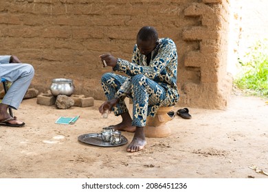Young African villager preparing tea sitting hunched over on a small footstool in his courtyard