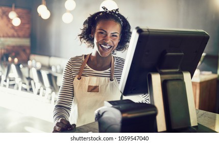 Young African waitress wearing an apron standing by a point of sale terminal and laughing while working in a trendy restaurant - Powered by Shutterstock