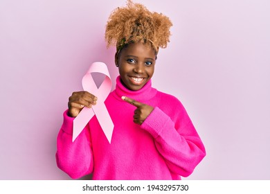 Young african woman with afro hair holding pink cancer ribbon smiling happy pointing with hand and finger  - Shutterstock ID 1943295730