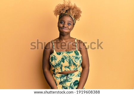 Young african woman with afro hair wearing summer dress puffing cheeks with funny face. mouth inflated with air, crazy expression. 
