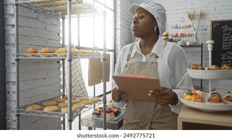 Young african woman in bakery room, holding a clipboard while inspecting baked goods on shelves inside the shop - Powered by Shutterstock