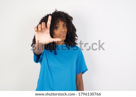 Young african woman with curly hair wearing casual blue shirt standing against white  background making fun of people with fingers on forehead doing loser gesture mocking and insulting.