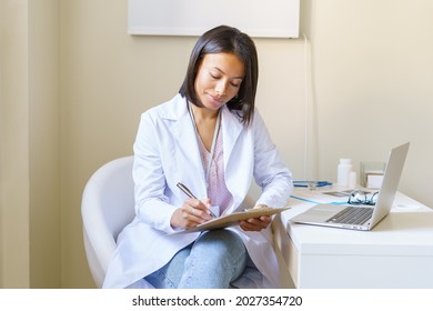 Young african woman doctor GP in white coat holding clipboard and filling patient form while working in medical clinic, afro american female medical worker doing paperwork at workplace with laptop