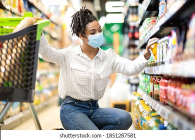 Young African Woman Doing Grocery Shopping Taking Food Product From Shelf In Supermarket Store Aisle, Wearing Protective Face Mask. Lady Customer Buying Groceries Essentials And Supplies Concept