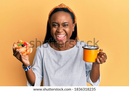 Young african woman drinking coffee and eating pastry celebrating crazy and amazed for success with open eyes screaming excited. 