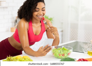 Young african woman eating salad at hone in kitchen, leaning on table and texting on smartphone, using an application - Shutterstock ID 1688555767