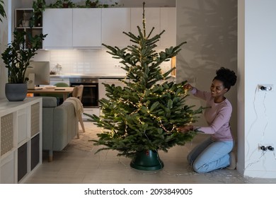 Young African Woman Enjoy Christmastime, Creating Minimalist Christmas Setting In House, Smiling Black Female Decorating Real Xmas Tree With Classic Led Lights, Creating Festive Mood At Home