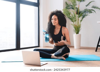 Young african woman fitness blogger vlogger trainer in sporty clothes drinking water while vlogging blogging online on laptop doing yoga class lesson remotely - Powered by Shutterstock