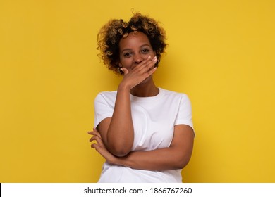 Young African Woman Giggles Covering Her Mouth With Hand, Hiding Her Emotion. Studio Shot On Yellow Wall.