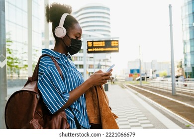 Young African Woman In Headphones And Facemask With Backpack Looking On Her Phone While Standing Outdoors