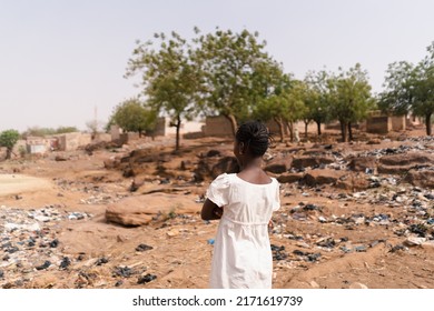Young African woman with her back to the camera looking at a polluted area full of windswept plastic bags and other items surrounding her village