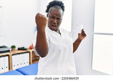 Young African Woman Holding Covid Record Card At Clinic Annoyed And Frustrated Shouting With Anger, Yelling Crazy With Anger And Hand Raised 