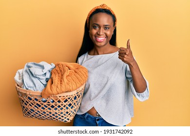 Young African Woman Holding Laundry Basket Smiling Happy And Positive, Thumb Up Doing Excellent And Approval Sign 