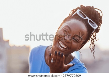 A young African woman laughs and blows a kiss. Close-up portrait of a charming dark-skinned smiling woman with sunglasses. An attractive girl is having fun on the background of the city.