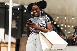 Young African Woman, In A Mall Using A Mobile Phone, With Bags Makes Purchases For Christmas, Shopping, Consumerism, Christmas