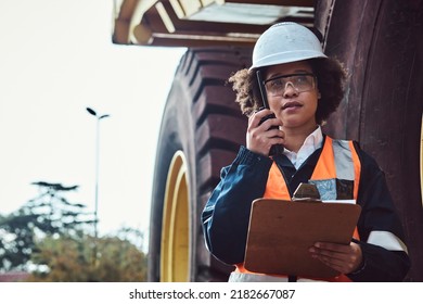 A young African woman mine worker is talking on a two way radio in front of a large haul dump truck wearing her personal protective wear
