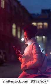 Young African Woman At Night With Smartphone Wait For Taxi From Booking Mobile App. Black Millennial Female Use Cellphone Outdoors Looking For Blind Date From Dating Application. Technology And Gadget