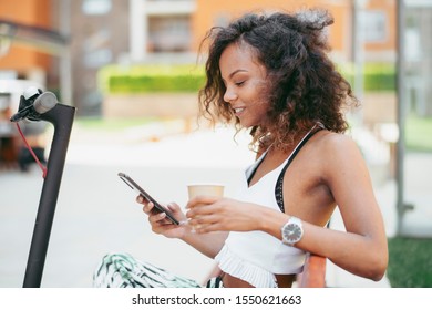 Young african woman outdoors. Beautiful woman sitting on bench drinking coffee and using phone. - Powered by Shutterstock