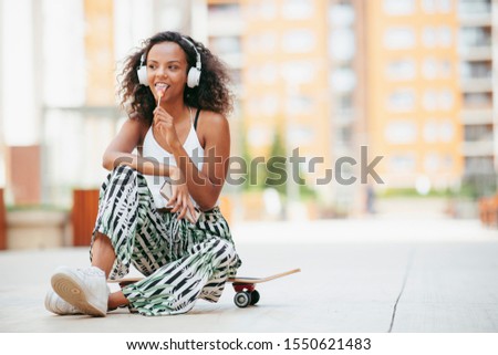 Young african woman outdoors. Beautiful woman sitting on skateboard and listening music.