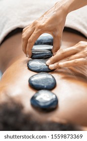 Young african woman with pebbles on her back on massage table in beauty spa salon. Closeup of woman masseuse hand placing lava stone on girl back. Lastone therapy at spa resort.