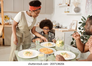 Young African woman putting plate with pasta on served table by her little son during family dinner - Powered by Shutterstock