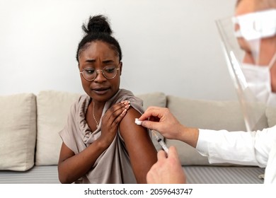 Young African Woman Receives A COVID-19 Vaccine Dose From A Male Healthcare Worker At Home. Male Doctor Giving A Covid-19 Vaccine To An African American Patient.