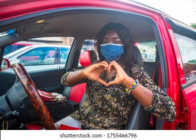 A Young African Woman Sitting In A Red Car, Wearing Face Mask For Protection And Showing A Love Sign Formed With Her Hands