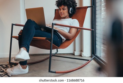 Young African Woman Sitting Relaxed On Chair Wearing Headphones And Working On Laptop. Female Software Programmer Working At Tech Startup.