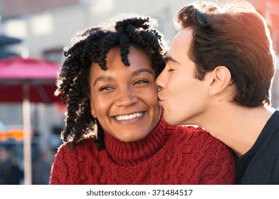 Young african woman is smiling while her boyfriend kissing her on the cheek. Close up face of young man kissing in woman's cheek while sitting at sidewalk cafe. Loving multi ethnic couple outdoor. - Shutterstock ID 371484517
