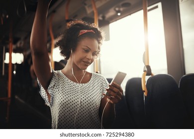 Young African woman smiling while standing by herself on a bus listening to music on a smartphone  - Powered by Shutterstock