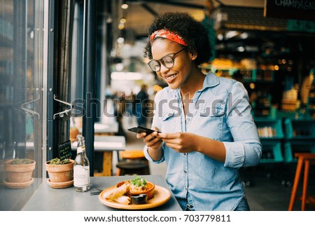 Young African woman smiling while sitting alone at a counter in a bistro taking photos of her meal with her smartphone