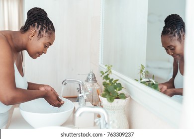 Young African woman standing over the sink in her bathroom washing her face with water in the morning