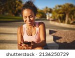 Young African woman in a tanktop and shorts smiling while sitting on steps in a park listening to music on her cellphone