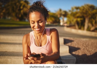 Young African woman in a tanktop and shorts smiling while sitting on steps in a park listening to music on her cellphone