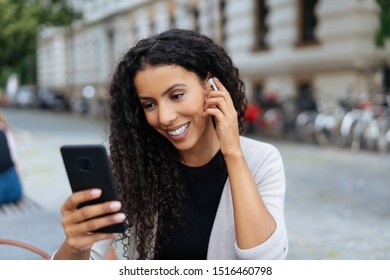 Young African woman using a ear bud with her mobile phone with a pleased smile in a quiet urban street - Powered by Shutterstock