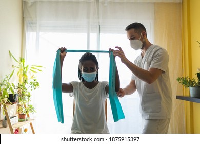 Young African Woman Wearing A Mask Doing Rehabilitation Exercises Of The Arms With A Rubber Band With A Physiotherapist Wearing A Mask And A Uniform In A Clinic