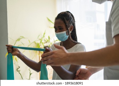 Young African Woman Wearing A Mask Doing Rehabilitation Exercises With A Rubber Band Next To A Physiotherapist Wearing A Mask And A Uniform In A Clinic