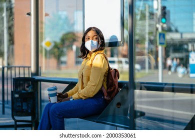 Young African Woman Wearing Protective Face Mask In A Tram Station Waiting For The Bus Alone. Black Female Student Sitting On Bench At Public Transportation Stop After Reopening Post Lockdown.