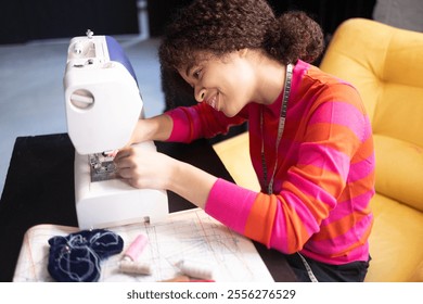 Young African woman working at sewing machine. Charming black girl in red sweater with measuring tape. Materials for sewing and fabric patterns in workplace. Seamstress is engaged in needlework - Powered by Shutterstock