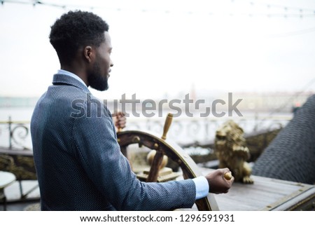 Young African-american businessman standing by sailing wheel and turning it while leading large ship