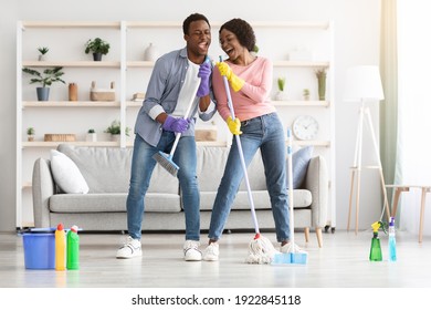 Young African-american Couple Cleaning Home, Playing With Mop And Broom, Having Fun In Living Room, Full Length Shot, Copy Space. Happy Black Family Singing And Dancing While Cleaning Apartment