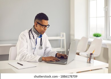 Young African-American doctor sitting at desk, using laptop computer, doing medical research on Internet, filling in electronic health records or giving consultation to patient on online eHealth chat - Powered by Shutterstock