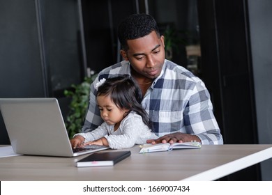 Young African-american Father A Designer At A Remote Work Works At A Computer Sits At A Table With His Charming Little Two-year-old Daughter. Concept Of Combining Work And Family